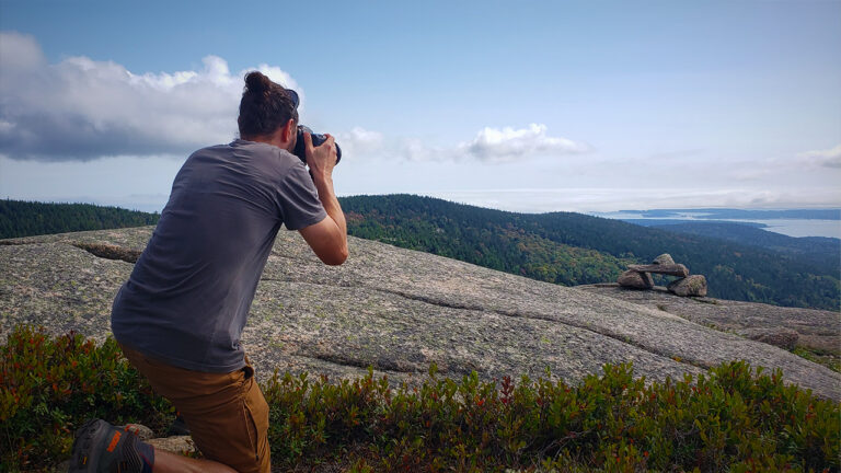 person kneeling on a mountain taking a photo of a mountain range