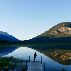 person standing on a dock in front of a lake mountains and open blue sky