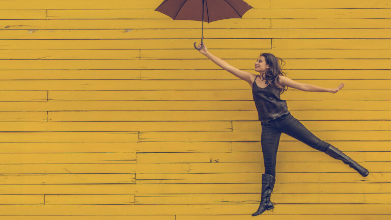 woman jumping in the air while holding a red umbrella against a yellow background